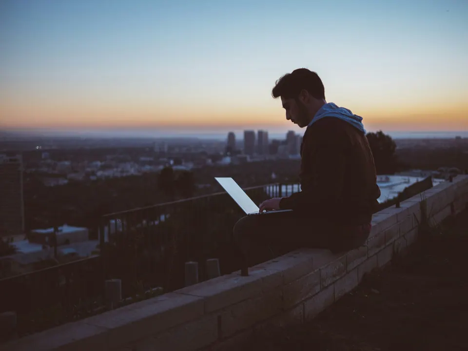 Homem sentado num muro com o notebook no colo, ao entardecer